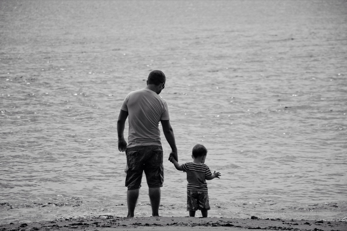 Fotografia realista: pai com o seu filho bebé ao colo à beira-mar, transmitindo serenidade e ternura